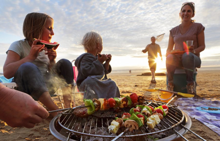 Una familia está gozando asando a la parilla kebabs juntos en la playa mientras que el sol comienza a fijar.